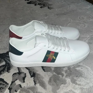 Gucci Ace modell - Säljer ett par vita Gucci sneakers i läder med klassiska gröna och röda ränder samt en guldfärgad bi på sidan. Baksidan har metallicdetaljer i rött och grönt med Gucci-logga. Snygg och stilren modell som sticker ut.