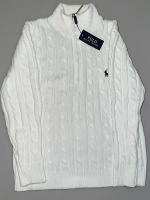 Vit kabelstickad tröja Polo Ralph Lauren - Vit kabelstickad tröja från Polo Ralph Lauren med halv dragkedja och hög krage. Tröjan har ribbade muddar och nederkant samt den klassiska broderade loggan på bröstet. Perfekt för en stilren och avslappnad look.
