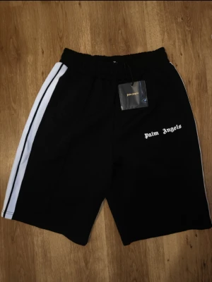 Svarta shorts från Palm Angels (förhandlingsbar) - Svarta shorts från Palm Angels med vita sidoränder och elastisk midja. Sportig look med logga tryckt på framsidan. Perfekta för en avslappnad stil eller träning. Materialet känns lätt och bekvämt. Helt ny .