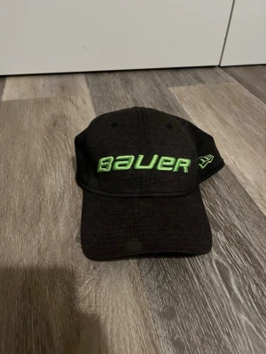 Bauer keps svart med grön logga - Svart sportkeps från Bauer med böjd skärm och broderad grön Bauer-logga framtill. Sidan har New Era-loggan i samma gröna färg. Kepsen är tillverkad i slitstarkt material och har klassisk form med justerbar baksida. Perfekt för träning och sport. Pris kan absolut diskuteras.