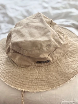 Beige bucket hat från Jacquemus - Säljer en beige bucket hat från  med bred brätte och råa kanter. Hatten är tillverkad i ett slitstarkt bomullstyg och har en diskret -logga framtill. Perfekt accessoar för en avslappnad och trendig look.