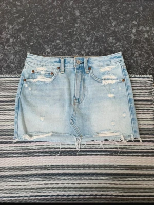 Ljusblå jeanskjol med slitningar - Trendig ljusblå jeanskjol i kort modell med slitna detaljer och fransig nederkant. Klassisk femficksdesign och bälteshällor. Perfekt för dig som gillar en avslappnad och cool stil.