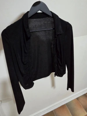 Svart cropped kofta med rynkade detaljer - Svart långärmad cropped kofta med rynkade detaljer framtill och klassisk krage. Tunn och mjuk i materialet, perfekt att slänga över en topp. Stängs med knappar framtill och har en stilren, minimalistisk look.