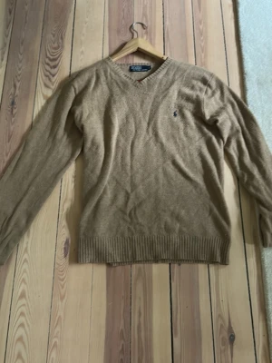 Beige stickad tröja Polo Ralph Lauren - Är dåligt på att ta bra bilder. Klassisk beige stickad tröja från Polo Ralph Lauren med V-ringning och diskret broderad logga på bröstet. Tröjan har långa ärmar och ribbade muddar vid ärmslut och nederkant. Perfekt för lager på lager och enkel att matcha.