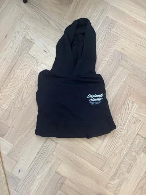 Svart hoodie Originals Studio - Svart hoodie från Originals Studio med vit logga tryckt på bröstet. Klassisk känguruficka framtill och stor huva. Tillverkad i mjukt bomullsmaterial som är skönt att ha på sig. Perfekt för en chill och avslappnad stil.