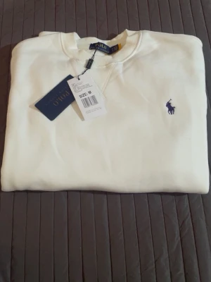 Vit sweatshirt från Polo Ralph Lauren - Klassisk vit sweatshirt från Polo Ralph Lauren med rund halsringning och broderad marinblå logga på bröstet. Tröjan har ribbade muddar vid ärmslut och nederkant. Perfekt för en clean och stilren look.
