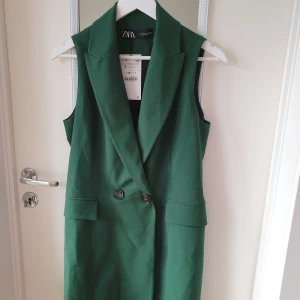 Grön ärmlös klänning från Zara - Snygg grön ärmlös klänning från Zara med dubbelknäppning framtill. Perfekt för en stilren look. Klänningen har en klassisk krage och fickdetaljer som ger en elegant touch. Ca 87cm lång