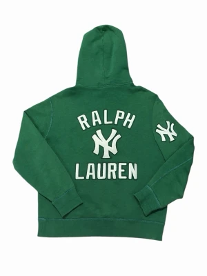 Grön Yankees hoodie från Ralph Lauren - Snygg grön hoodie från Ralph Lauren med Yankees-tryck på bröstet och ärmen. Stor broderad logga på ryggen och klassisk huva med dragsko. Mjuk bomullsblandning och känguruficka framtill. Perfekt för dig som gillar sportig streetstyle.