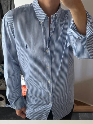 Blårandig skjorta från Ralph Lauren - Klassisk blårandig skjorta från Ralph Lauren med button-down krage och broderad logga på bröstet. Skjortan har långa ärmar, vita knappar och är tillverkad i mjuk bomull. Perfekt för en clean och stilren look.