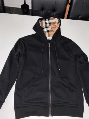 Burrbery hoodie - Snygg svart hoodie från Burberry med dragkedja framtill och klassiskt rutmönstrat foder i huvan. Hoodien har två stora fickor fram och justerbar dragsko i huvan. Perfekt för dig som gillar stilrena plagg med exklusiva detaljer.