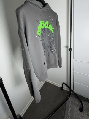 Sp5der hoodie - Säljer en grå hoodie från Sp5der med stor neongrön logga och spindelnätsmönster på bröstet. Hoodien har luva, ribbade muddar och en avslappnad passform. Materialet känns som mjuk bomullsblandning och trycket har coola detaljer i vitt och svart.