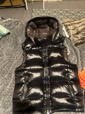 Moncler vest  - Snygg svart dunväst från Moncler med glansig finish och stor huva. Västen har två dragkedjeförsedda fickor framtill och klassisk Moncler-logga på bröstet. Perfekt för lager-på-lager och streetwear-stil.
