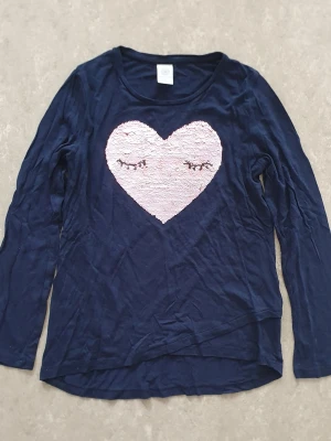 Tröja - Marinblå långärmad tröja med ett stort rosa hjärta i paljetter på bröstet. Hjärtat har broderade ögonfransar för en söt och unik look. Tröjan har rund halsringning och är gjord i ett mjukt, tunt material.