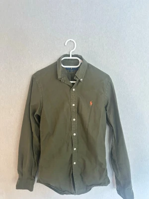 Olivgrön linneskjorta Ralph Lauren  - Snygg olivgrön skjorta från Ralph Lauren i slim fit-modell. Tillverkad i linne med klassisk krage, knappar framtill och den ikoniska broderade loggan på bröstet. Perfekt för en clean och stilren look. Använd fåtal gånger, schyssta priser vid snabb affär.
