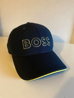 Mörkblå keps från BOSS med gul detalj - Snygg mörkblå keps från BOSS med broderad logga i gult framtill. Klassisk böjd skärm med gul kant och justerbar baksida. Tillverkad i slitstark bomull som ger en sportig och clean look.