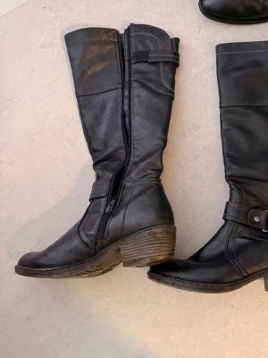 Svarta höga boots med klack - Säljer ett par svarta höga stövlar med grov klack och dekorativt spänne vid ankeln. Dragkedja på insidan för enkel på- och avtagning. Tillverkade i ett slitstarkt syntetmaterial med matt finish och markerade sömmar.