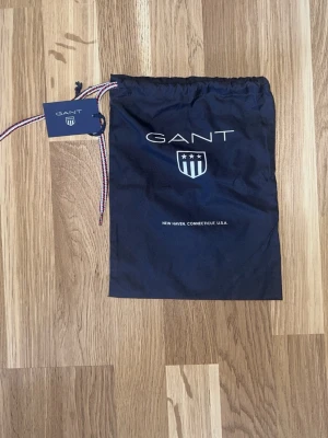 Marinblå påse från GANT - Snygg marinblå tygpåse från GANT med logga och emblem framtill. Påsen har dragsko upptill med ett band i rött, vitt och blått samt en etikett med GANT-loggan. Perfekt för att förvara accessoarer eller mindre prylar.