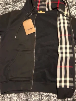 Svart Burberry hoodie med dragkedja - Svart hoodie från Burberry med klassiskt rutigt foder i huvan och längs dragkedjan. Hoodien har fickor framtill, ribbade muddar och justerbar huva med snören. Perfekt för dig som gillar stilrena plagg med en touch av lyx.
