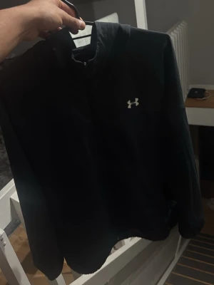 Svart hoodie från Under Armour - Svart hoodie från Under Armour med dragkedja och huva. Logotypen är tryckt på bröstet och texten UNDER ARMOUR finns längs ena ärmen. Materialet är mjukt och bekvämt, perfekt för en sportig look. Passar dig som gillar stilrena och funktionella kläder.
