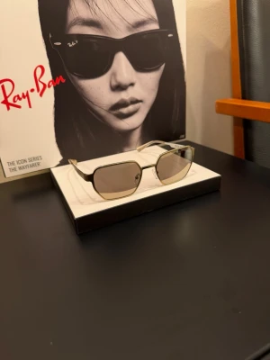 Rektangulära solglasögon Michael Kors - Snygga solglasögon från Michael Kors med rektangulära linser och tunna guldfärgade bågar. Glasen är gråtonade och bågarna har en elegant och modern design med diskret logga på sidan. Perfekta för dig som vill ha en stilren accessoar.