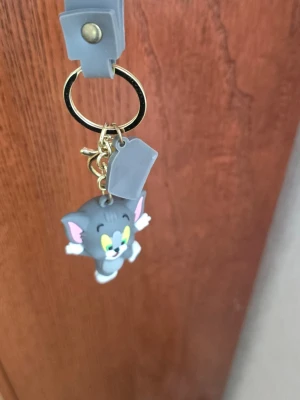 Tom & Jerry nyckelring grå - Säljer en grå nyckelring med Tom från Tom & Jerry i mjukt gummi. Nyckelringen har guldfärgad ring och grå rem med Jerry-tryck. Perfekt accessoar för dig som gillar tecknade klassiker och vill ha något unikt på väskan eller nycklarna.
