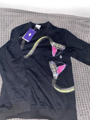 Svart sweatshirt med kobramotiv - Cool svart sweatshirt från Daily Paper med unikt tryck av två kobror med rosa detaljer på framsidan. Tröjan har rund hals, ribbade muddar vid ärmslut och nederkant samt en mjuk bomullskänsla. Perfekt för dig som vill sticka ut med en edgy streetwear-look.