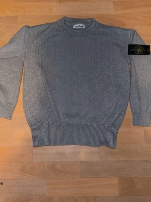 Grå stickad tröja från Stone Island - Snygg grå stickad tröja från Stone Island med klassisk rund hals och ribbade muddar. Tröjan har den ikoniska svarta patchen med gul och grön logga på vänster ärm. Perfekt för dig som gillar streetwear och vill ha något stilrent.
