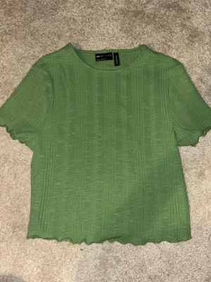 Grön ribbad topp från ASOS DESIGN - Snygg grön ribbad topp från ASOS DESIGN med korta ärmar och vågig kant nertill. Toppen har en enkel rund halsringning och är tillverkad i ett mjukt material som känns skönt mot huden. Perfekt att matcha med jeans eller kjol för en trendig look. Den är absolut xs/s