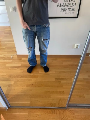 Blå slitna diesel jeans - Säljer ett par blå diesel jeans med avslappnad passform och stora slitningar på båda benen. Jeansen har klassisk femficksdesign och raka ben. Perfekta för en streetwear-look och passar dig som gillar en edgy stil.