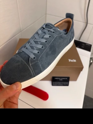 Blå mocka sneakers från Louboutin - Snygga blå sneakers i mocka från Christian Louboutin med klassisk röd sula och vita kanter. Skorna har rund tå, matchande blå skosnören och diskret branding på insidan. Perfekta för dig som vill ha en stilren och exklusiv look.