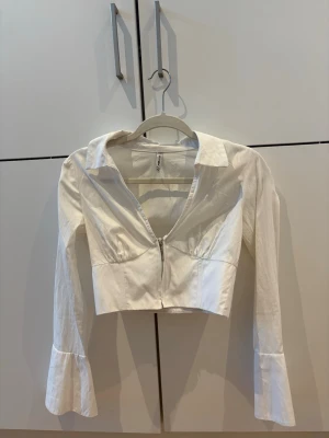 Vit croppad skjorta med dragkedja - Säljer en vit croppad blus med lång ärm och vida ärmar. Toppen har en djup v-ringning och dragkedja framtill. 