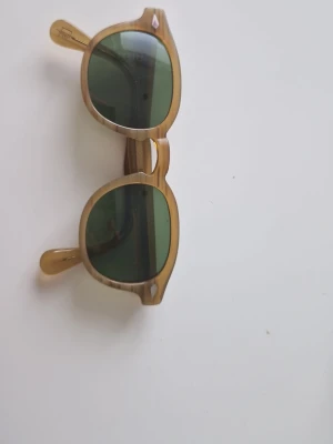 Bruna solglasögon från Moscot - Säljer ett par snygga solglasögon från Moscot med rundad form och transparent brun båge. Glasen är mörkgröna och bågarna har en cool retrostil. Perfekta för dig som vill sticka ut med en klassisk NYC-look.
