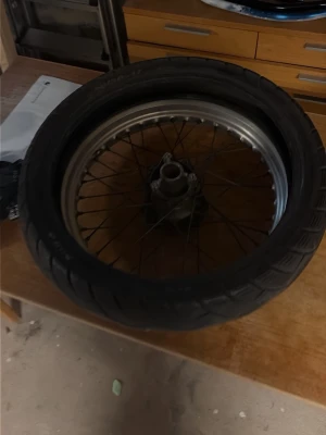 Svart motorcykeldäck med fälg - Säljer ett svart motorcykeldäck med silverfärgad metallfälg och ekrar. Däcket har ett tydligt mönster och fälgen har nav i mitten. Perfekt för dig som behöver ett extra hjul till din hoj.