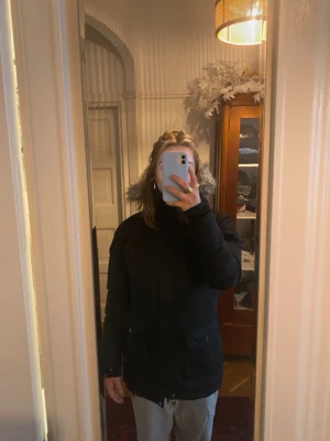 Svart parkas med fuskpäls från Everest - Snygg svart parkas från Everest med avtagbar huva och fuskpäls. Jackan har två stora fickor framtill, dragkedja och knappar, samt ribbade muddar vid ärmsluten. Perfekt för kalla dagar och riktigt skön passform.