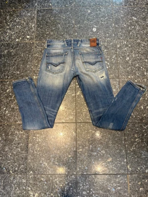 Blå Replay jeans med slitningar - Säljer ett par blå jeans från Replay med snygga slitningar och tvättade detaljer. Klassisk femficksmodell med raka ben och läderpatch bak i midjan. Jeansen är tillverkade i bomull och har coola sömmar på bakfickorna.