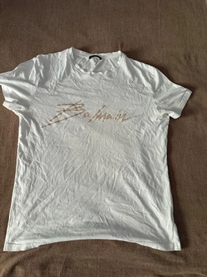 Vit Balmain t-shirt med logga - Snygg vit t-shirt från Balmain med beige logga i coolt typsnitt över bröstet. Klassisk rund halsringning och korta ärmar. Perfekt för dig som gillar stilrena och trendiga plagg med exklusiv känsla. Tillverkad i mjuk bomull för skön komfort.