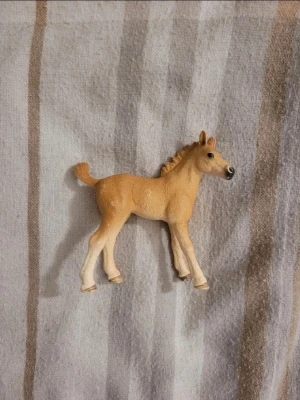 Liten plastfigur av föl - Säljer en liten plastfigur som föreställer ett ljusbrunt föl med beige ben och mörka ögon. Figuren har fina detaljer och är perfekt för dig som gillar djur eller samlar på hästfigurer.