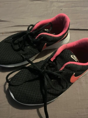 Svarta Nike sneakers med rosa detaljer - Säljer ett par svarta Nike sneakers med rosa swoosh och rosa insida. Skorna har vit sula och är tillverkade i meshmaterial som andas. Klassisk låg modell med snörning och sportig look. Perfekta för dig som gillar stilrena och bekväma sneakers. Stl 36. Kan tänka mig att sänka priset skriv bara.