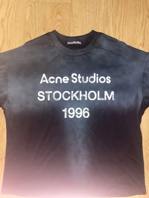 Svart t-shirt Acne Studios Stockholm - Cool svart t-shirt från Acne Studios med vit text 'Acne Studios STOCKHOLM 1996' på bröstet. Klassisk rund hals och korta ärmar. Perfekt för dig som gillar streetwear och vill ha en stilren look.
