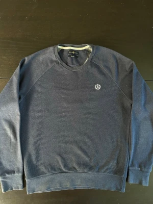 Mörkblå sweatshirt Henri Lloyd - Snygg mörkblå  sweatshirt från Henri Lloyd med rund halsringning och diskret logga broderad på bröstet. Tröjan har en strukturerad stickning framtill och släta ärmar samt muddar vid ärmslut och nederkant. Perfekt för en stilren och avslappnad look.