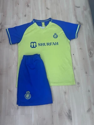 Gul och blå fotbollströja med shorts - Säljer ett matchande set med gul fotbollströja och blå shorts från SHURFAH. Tröjan har blå ärmar och detaljer samt tryckt logga och text på bröstet. Shortsen är helblå med logga. Perfekt för träning eller match!