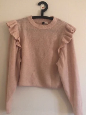 Ljusrosa stickad tröja med volanger - Supersöt ljusrosa stickad tröja från H&M med volangdetaljer längs axlarna. Tröjan har rund halsringning, långa ärmar och är gjord i ett mjukt, fluffigt material. Perfekt för dig som gillar en feminin och trendig stil.