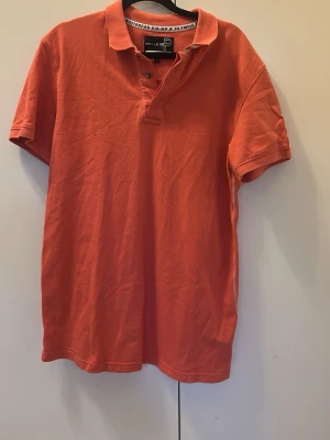 Orange pikétröja från Pelle P - Säljer en orange pikétröja från Pelle P i klassisk modell med krage och knappar framtill. Tröjan är tillverkad i bomull och har korta ärmar. Perfekt för en avslappnad och sportig look.