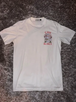Vit t-shirt från Casablanca - Vit t-shirt från Casablanca med tryck på bröstet som visar en båge och texten 'Casa Blanca' i rött. Klassisk rund hals och korta ärmar. Tillverkad i mjuk bomull för en skön känsla och clean look. Sitter som en M