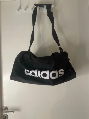 Svart sportbag från Adidas - Snygg svart sportbag från Adidas med stort vitt logotryck på sidan. Väskan har justerbar axelrem och bärhandtag, perfekt för gymmet eller träning. Tillverkad i slitstarkt tygmaterial och har dragkedja upptill för enkel åtkomst.