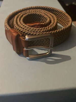 Flätat beige bälte med skinn-detaljer - Snyggt flätat bälte i beige med bruna skinn-detaljer och silverfärgat spänne. Perfekt accessoar för att lyfta din outfit med en stilren touch. Passar till både jeans och chinos.