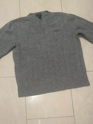 Grå stickad tröja från GANT - Mysig grå stickad tröja från GANT med V-ringning och diskret logga broderad på bröstet. Tröjan har långa ärmar och ribbade muddar vid ärmslut och nederkant. Perfekt för lager på lager och klassisk stil.
