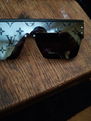 Louis Vuitton svarta solglasögon - Säljer ett par statement solglasögon från Louis Vuitton med spegelglas och ikoniskt LV-mönster över hela linsen. Glasögonen har en futuristisk, rektangulär form och svarta skalmar. Perfekta för dig som vill sticka ut och addera lyx till din stil. De är i bra skick.