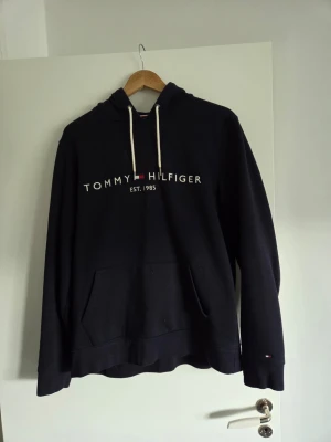 Mörkblå hoodie Tommy Hilfiger - Snygg mörkblå hoodie från Tommy Hilfiger med vit dragsko i huvan och klassiska loggan broderat på bröstet. Stor känguruficka framtill och liten logga på ärmen. Tillverkad i mjukt bomullsmaterial, perfekt för en avslappnad stil.