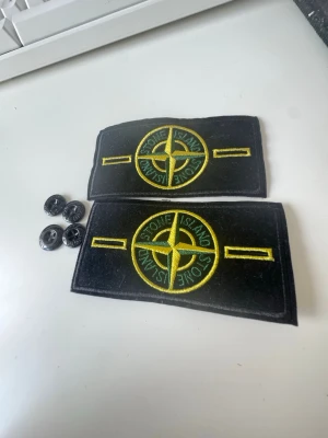 Stone Island badge bundle - Två svarta tygmärken från Stone Island med det klassiska gula och gröna kompasslogotypen broderad framtill. Märkena kommer med fyra svarta knappar för enkel fastsättning på plagg. Perfekt för att ge din jacka eller tröja en unik touch.